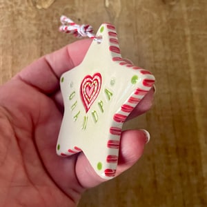 Puede incluir: Adorno de cer&aacute;mica en forma de estrella con borde a rayas rojas y blancas y dise&ntilde;o de coraz&oacute;n rojo. El adorno tiene las palabras "GIA NDPA" en verde. Se adjunta una cuerda roja, blanca y verde para colgar.