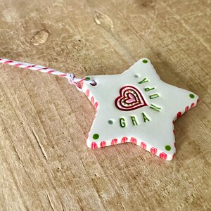 Puede incluir: Adorno de cer&aacute;mica blanco en forma de estrella con un dise&ntilde;o de coraz&oacute;n rojo y rosa y la palabra "GRA" en verde. El adorno tiene un borde a rayas rojo y rosa y puntos verdes. Se adjunta una cuerda roja y blanca.