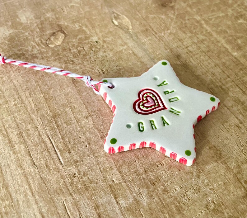 Puede incluir: Adorno de cer&aacute;mica blanco en forma de estrella con un dise&ntilde;o de coraz&oacute;n rojo y rosa y la palabra "GRA" en verde. El adorno tiene un borde a rayas rojo y rosa y puntos verdes. Se adjunta una cuerda roja y blanca.