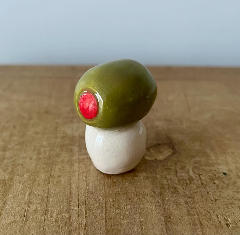 Pu&ograve; includere: Una scultura in ceramica a forma di oliva verde con dettaglio di peperone rosso, su una base bianca. Un oggetto decorativo stravagante.