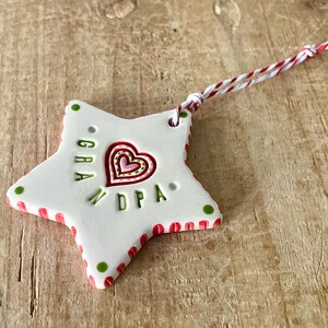 Puede incluir: Adorno blanco en forma de estrella con detalles rojos y verdes. El adorno presenta un dise&ntilde;o de coraz&oacute;n y las letras "GRA" y "DPA". Una cuerda roja y blanca est&aacute; unida para colgar. El adorno est&aacute; sobre una superficie de madera.