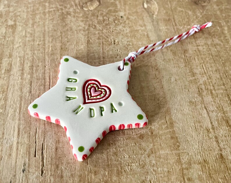Puede incluir: Adorno blanco en forma de estrella con detalles rojos y verdes. El adorno presenta un dise&ntilde;o de coraz&oacute;n y las letras "GRA" y "DPA". Una cuerda roja y blanca est&aacute; unida para colgar. El adorno est&aacute; sobre una superficie de madera.