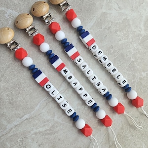 May include: Four dummy clips featuring wooden beads at the top, followed by red, white, and blue silicone beads. Each clip includes a cube bead with the French flag design and letter beads spelling out the names OLISE, MBAPPE, SALIBA, and DEMBELE.