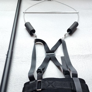May include: A black exercise device with a long, adjustable pole, a metal frame, and a harness. The harness has a padded waist belt and shoulder straps. The device is designed for fitness and strength training.