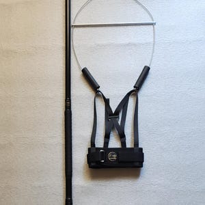 May include: A black training aid for horses, featuring a long, textured pole, a circular frame, and a harness-like structure. The harness has adjustable straps and a buckle. The frame is connected to the pole by a wire.