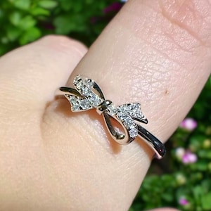 Puede incluir: Un anillo de plata con un pequeño diseño de lazo de diamantes brillantes. El anillo se lleva en un dedo.