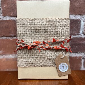 Puede incluir: Una caja de regalo beige envuelta con arpillera y adornada con una guirnalda decorativa de hojas naranjas. Una pequeña etiqueta con un sello de signo de interrogación cuelga de la caja, lo que sugiere un regalo misterioso.