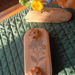 May include: A rectangular light-colored wooden block featuring a printed gray floral design and two star-shaped wooden knobs. Behind it, a piece of driftwood holds four glass flowers in green, yellow, blue, and orange colors.