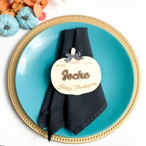 May include: A Thanksgiving place setting with a teal plate and a black napkin. A wooden pumpkin-shaped place card with the name "Joche" and the words "Happy Thanksgiving" is on the napkin. The pumpkin has a black and white checkered bow.