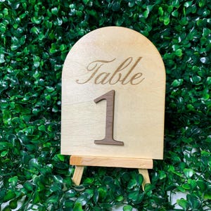May include: A wooden table number sign with the word "Table" and the number "1" engraved on it. The sign is light-colored and stands on a small wooden easel. The background is a dense arrangement of green leaves.