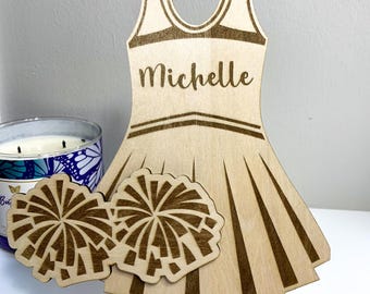 May include: Wooden cutout of a cheerleader uniform with the name "Michelle" on the front. The uniform is brown and has white stripes. The cutout is on a small wooden easel and includes two pom-poms.