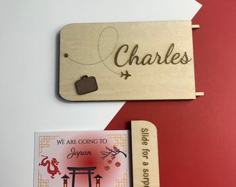 May include: A wooden nameplate engraved with the name "Charles", a dotted line, a suitcase, and an airplane. A card below reads "We are going to Japan" with Japanese imagery. A wooden slider reads "Slide for a surprise".