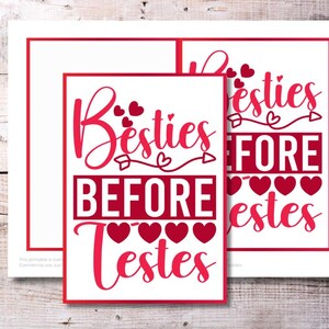 May include: Two white prints with red borders and text on a wooden background. The prints say "Besties Before Testes" in a cursive font with heart accents. The prints are a Valentine's Day or friendship theme.