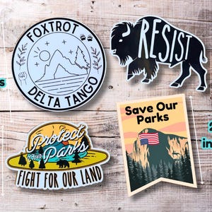 May include: Four vinyl stickers with different designs. The first sticker is blue and white with the text "FOXTROT DELTA TANGO". The second sticker is black and white with the text "RESIST". The third sticker is yellow and green with the text "Protect Our Parks Fight For Our Land". The fourth sticker is orange and green with the text "Save Our Parks".