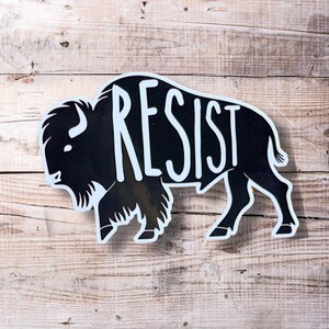 May include: Black and white silhouette of a buffalo with the word "RESIST" written across its body. The buffalo is on a woodgrain background.