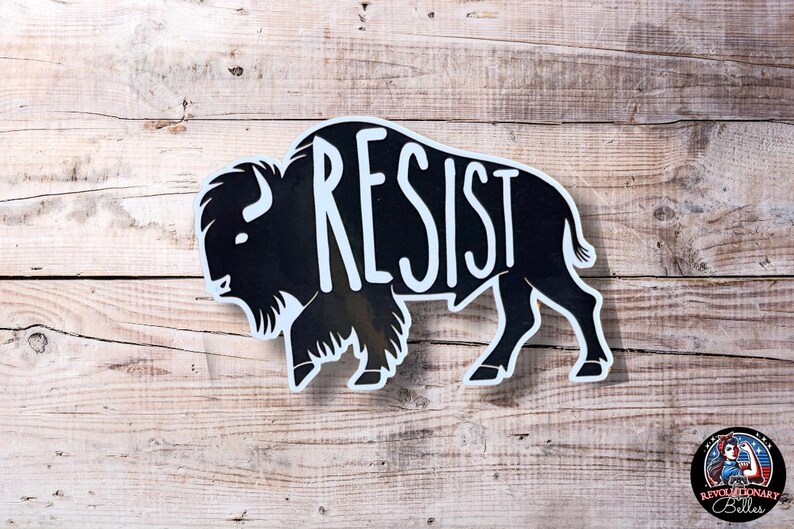 May include: Black and white silhouette of a buffalo with the word "RESIST" written across its body. The buffalo is on a woodgrain background.