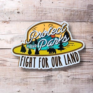 May include: A white sticker with a yellow sun and blue lake design. The text reads "Protect Our Parks" and "Fight For Our Land".
