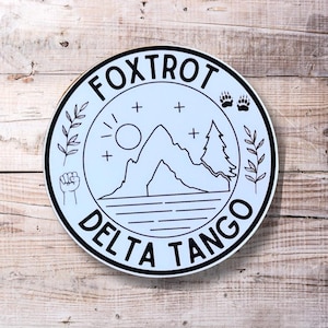 May include: A white circle sticker with a black outline and the text "FOXTROT DELTA TANGO" around the edge. Inside the circle is a black and white illustration of a mountain, a sun, two trees, and a fist.