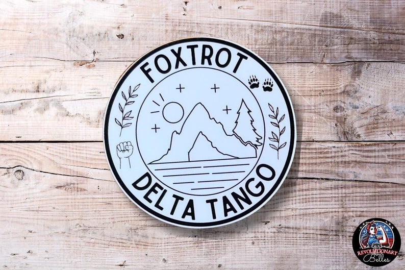 May include: A white circle sticker with a black outline and the text "FOXTROT DELTA TANGO" around the edge. Inside the circle is a black and white illustration of a mountain, a sun, two trees, and a fist.
