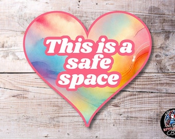 Puede incluir: Una pegatina en forma de corazón rosa con un fondo de acuarela. La pegatina dice "This is a safe space" en letras blancas.