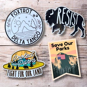 May include: Five different stickers with various designs. The stickers are on a wooden background. The stickers include a black and white mountain scene with the text "FOXTROT DELTA TANGO", a black silhouette of a buffalo with the text "RESIST", a yellow and green sticker with a mountain scene and the text "Protect Our Parks Fight For Our Land", a yellow and green sticker with a mountain scene and the text "Save Our Parks", and a sticker with a woman holding a flag and the text "Revolutionary Belles".