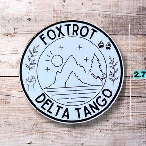 Puede incluir: Una pegatina redonda blanca y negra con el texto "FOXTROT DELTA TANGO" en un c&iacute;rculo. La pegatina presenta una monta&ntilde;a, un sol, dos &aacute;rboles y un pu&ntilde;o. La pegatina tiene un di&aacute;metro de 2,75 pulgadas.