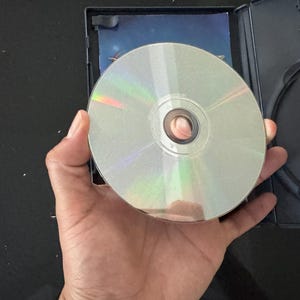 May include: A silver-colored disc with a central hole, likely a CD or DVD, is held in a hand against a black background.