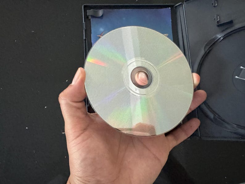 May include: A silver-colored disc with a central hole, likely a CD or DVD, is held in a hand against a black background.