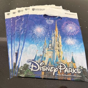 May include: Several Disney Parks shopping bags. Each bag displays a castle scene with fireworks against a blue backdrop. The Disney Parks logo is visible. The bags are made of recycled plastic.