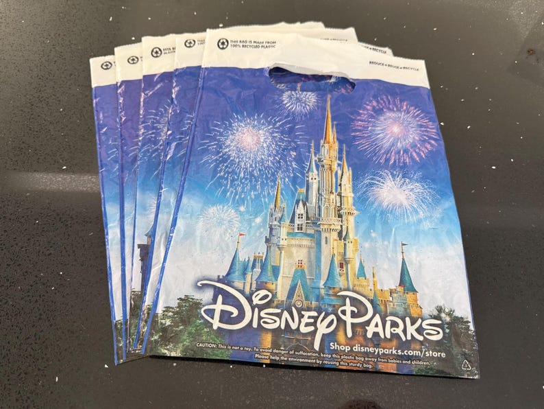 May include: Several Disney Parks shopping bags. Each bag displays a castle scene with fireworks against a blue backdrop. The Disney Parks logo is visible. The bags are made of recycled plastic.