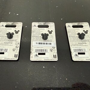 May include: Three Disney pins with a black Mickey Mouse head design. Each pin is attached to a white backing card with printed text, including the Disney Store logo and a barcode. The pins are displayed on a dark, reflective surface.