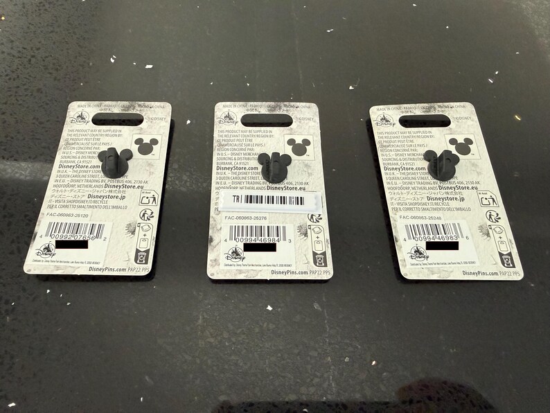 May include: Three Disney pins with a black Mickey Mouse head design. Each pin is attached to a white backing card with printed text, including the Disney Store logo and a barcode. The pins are displayed on a dark, reflective surface.