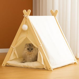 May include: A wooden cat bed in the shape of a teepee, with a cream canvas cover and a cream cushion. A fluffy grey cat sits inside. A white pom-pom hangs from the top. The bed is designed for indoor use.