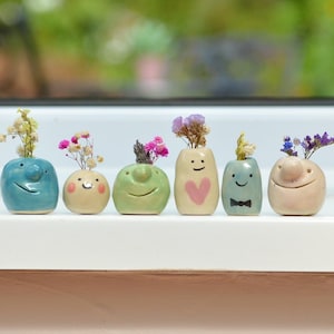 May include: Six small ceramic vases with faces, each holding dried flowers. The vases are painted in various colours, including blue, green, yellow, pink, and white.