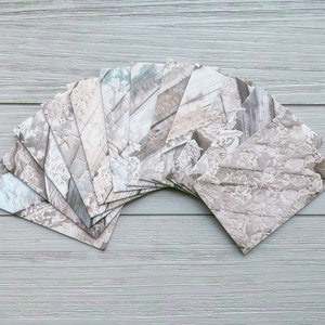 May include: A collection of rectangular paper envelopes arranged in a fan shape. Each envelope features a printed pattern of white lace overlaying a light grey and beige wood grain texture. The envelopes are uniform in size and shape.