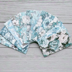May include: A set of rectangular paper envelopes arranged in a fan shape, featuring a repeating floral pattern in shades of teal, blue, and white. The envelopes display detailed illustrations of various blooming flowers and leaves across their surfaces.