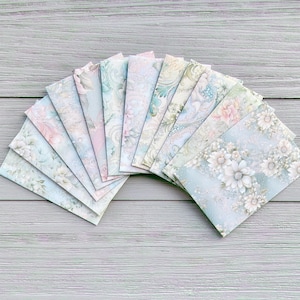 Op de afbeelding: Een waaier van 12 delicate, met bloemen bedrukte papieren enveloppen in tinten blauw, groen en roze. De enveloppen zijn in een halve cirkel gerangschikt met de punten van de enveloppen naar buiten gericht.