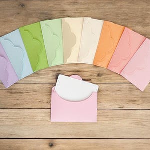 Puede incluir: Una colección de sobres coloridos con bordes festoneados en tonos pastel, dispuestos en arco sobre una superficie de madera. Un sobre rosa contiene una tarjeta blanca. Los sobres son en tonos lavanda, azul, verde, amarillo y naranja.