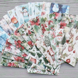 May include: A collection of rectangular paper sheets featuring various Christmas-themed illustrations, including snowmen, poinsettias, pine trees, red ornaments, and winter landscapes, arranged in a fan-like pattern on a gray wooden surface.