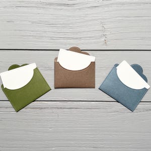 May include: Three small, square-shaped card holders in olive green, brown, and slate blue. Each holder features a curved flap opening and contains a plain white rectangular card tucked inside, displaying a clean, simple design for organizing small items.