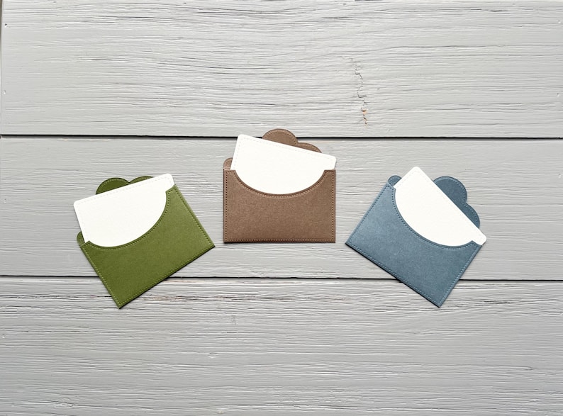May include: Three small, square-shaped card holders in olive green, brown, and slate blue. Each holder features a curved flap opening and contains a plain white rectangular card tucked inside, displaying a clean, simple design for organizing small items.