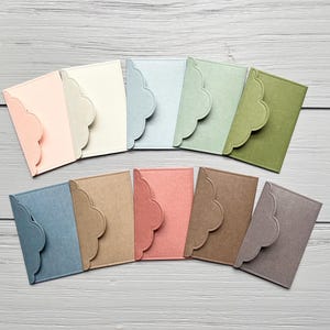 May include: A collection of eleven rectangular card holders or envelopes in various colors including light pink, cream, light blue, sage green, olive green, slate blue, tan, coral, brown, and grey, each featuring a scalloped edge flap closure.
