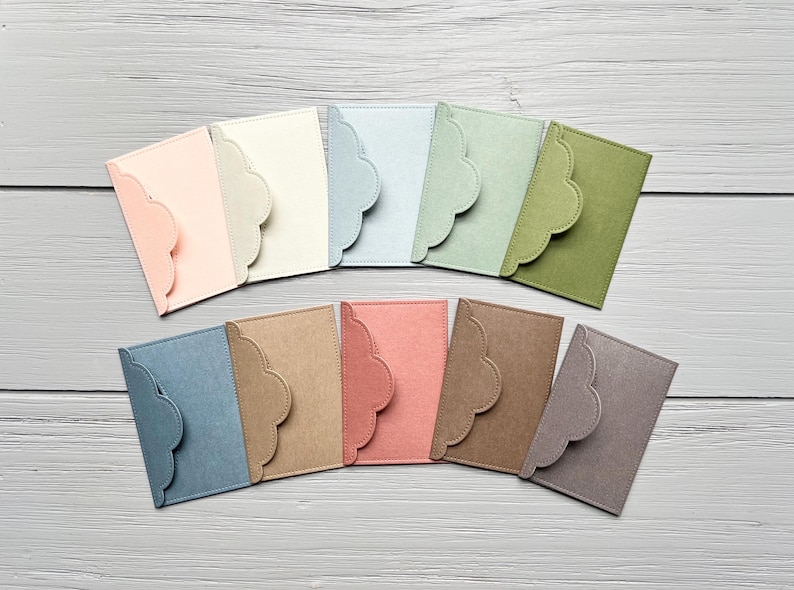 May include: A collection of eleven rectangular card holders or envelopes in various colors including light pink, cream, light blue, sage green, olive green, slate blue, tan, coral, brown, and grey, each featuring a scalloped edge flap closure.