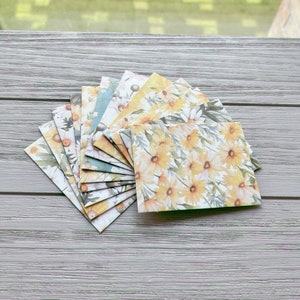May include: A fan-shaped arrangement of rectangular paper cards featuring a repeating watercolour-style pattern of yellow daisies and green foliage. The cards are layered on top of one another, displaying various shades of yellow, white, and green.