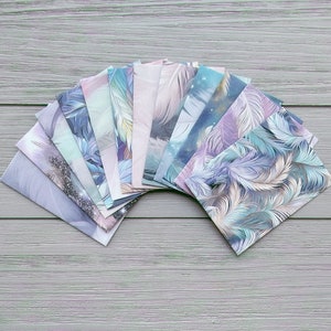 May include: A collection of rectangular cards arranged in a fan shape, each featuring distinct printed designs. The patterns include various depictions of feathers in shades of blue, purple, teal, and white, with some cards showing glittery accents.