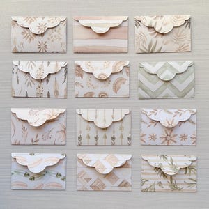 Könnte beinhalten: Eine Sammlung von zwölf rechteckigen Papierumschlägen mit gewellten Klappen, die jeweils ein einzigartiges Muster in neutralen Beige-, Creme- und sanften Grüntönen aufweisen, darunter Designs mit Federn, Blättern, Pfeilen, Monden und Streifen.