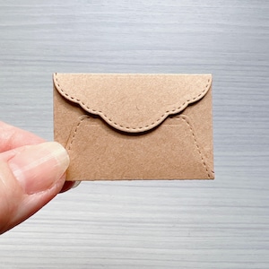 May include: A small, rectangular, light brown kraft paper envelope featuring a scalloped flap edge and decorative embossed stitching detail along the perimeter of the flap and the main body of the envelope, creating a textured appearance.