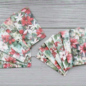 May include: A set of rectangular fabric napkins featuring a festive botanical print with red poinsettia flowers, green leaves, and small red berries. The napkins are arranged in two fanned stacks, displaying the repeating floral pattern throughout.
