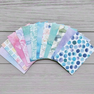 May include: A collection of patterned paper sheets in pastel shades. The designs feature watercolour washes, geometric shapes, and floral patterns. The papers are arranged on a grey wooden surface, displaying a variety of artistic designs.