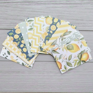 May include: A collection of rectangular paper cards arranged in a fan shape, featuring various patterns including yellow lemons, green leaves, white blossoms, yellow chevron stripes, white polka dots, and geometric shapes in shades of yellow, blue, and white.